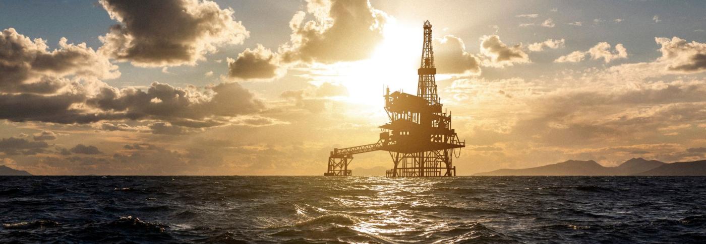 An offshore oil rig silhouetted against a dramatic sunset sky with scattered clouds, surrounded by ocean waves.