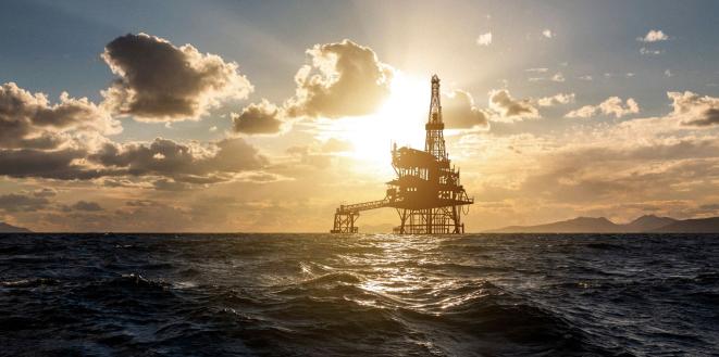 An offshore oil rig silhouetted against a dramatic sunset sky with scattered clouds, surrounded by ocean waves.