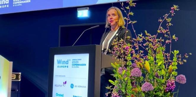 A lady stands at a podium giving a presentation at the WindEurope Annual Event 2025 in Copenhagen. The podium displays the event logo and names of sponsors. In the foreground is a large floral arrangement with purple and yellow flowers. A blue backdrop with event branding is visible behind the speaker.